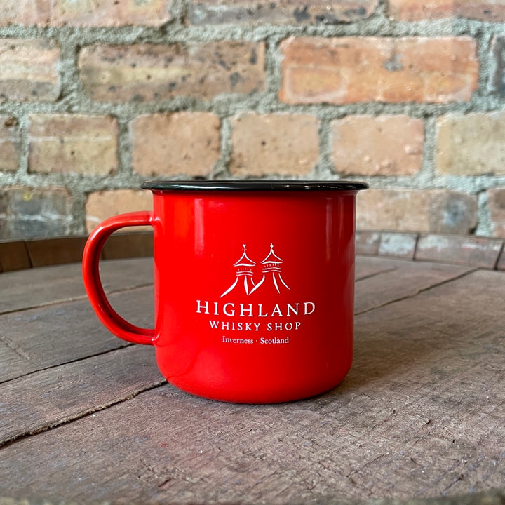 Red enamel mug with 'Highland Whisky Shop' branding on a wooden surface with a brick wall background.