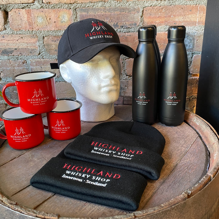Highland Whisky Shop branded merchandise including a cap, mugs, bottles, and a towel on a wooden barrel with a brick background.
